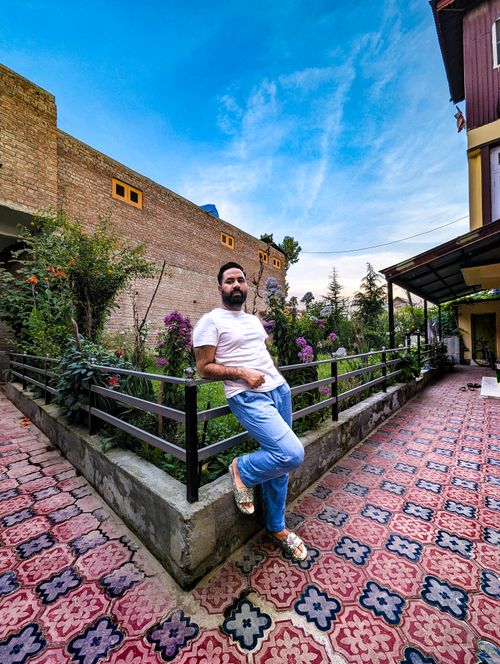 Free A man poses casually in a garden, leaning on a railing, wearing a white t-shirt and jeans. Stock Photo