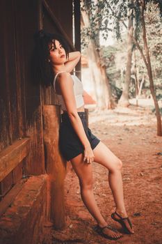 Caucasian woman in a white tank top and black shorts posing against a wooden wall.