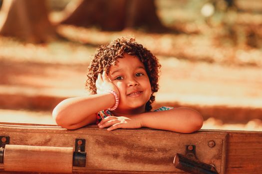 A cheerful young girl with curly hair resting on a bench in a sunny park.