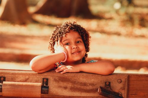 Free A cheerful young girl with curly hair resting on a bench in a sunny park. Stock Photo
