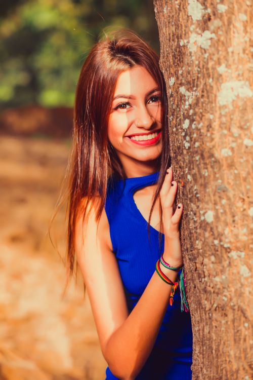 Free Portrait of a smiling woman in a blue dress leaning on a tree outdoors, showcasing natural beauty. Stock Photo