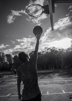 A dramatic black and white silhouette of a man reaching up to shoot a basketball outdoors.