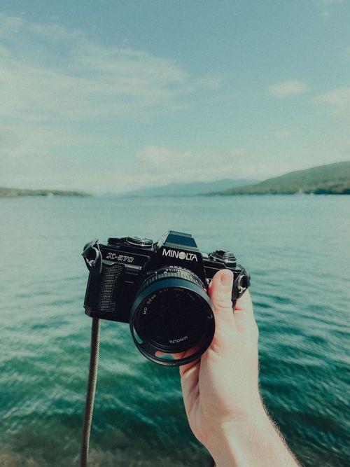 Free Hand holding a vintage camera against the background of a tranquil blue lake under a clear sky. Stock Photo