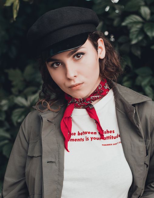 Portrait of a young woman wearing a black hat and red scarf outdoors with foliage background.