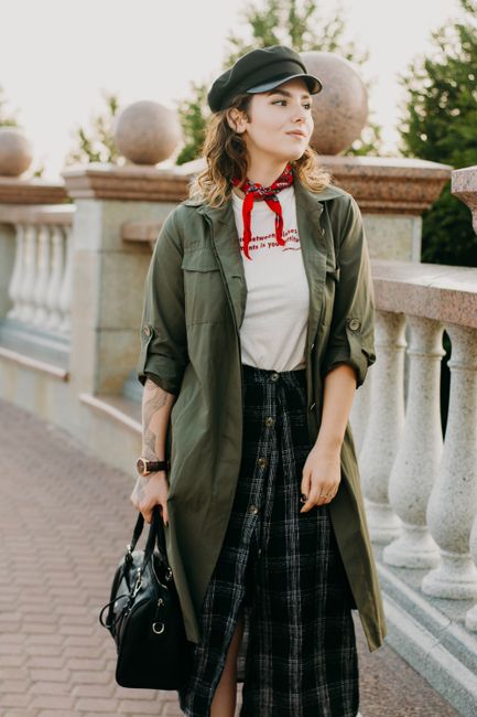 Fashionable woman in green jacket and plaid skirt posing outdoors with black bag.