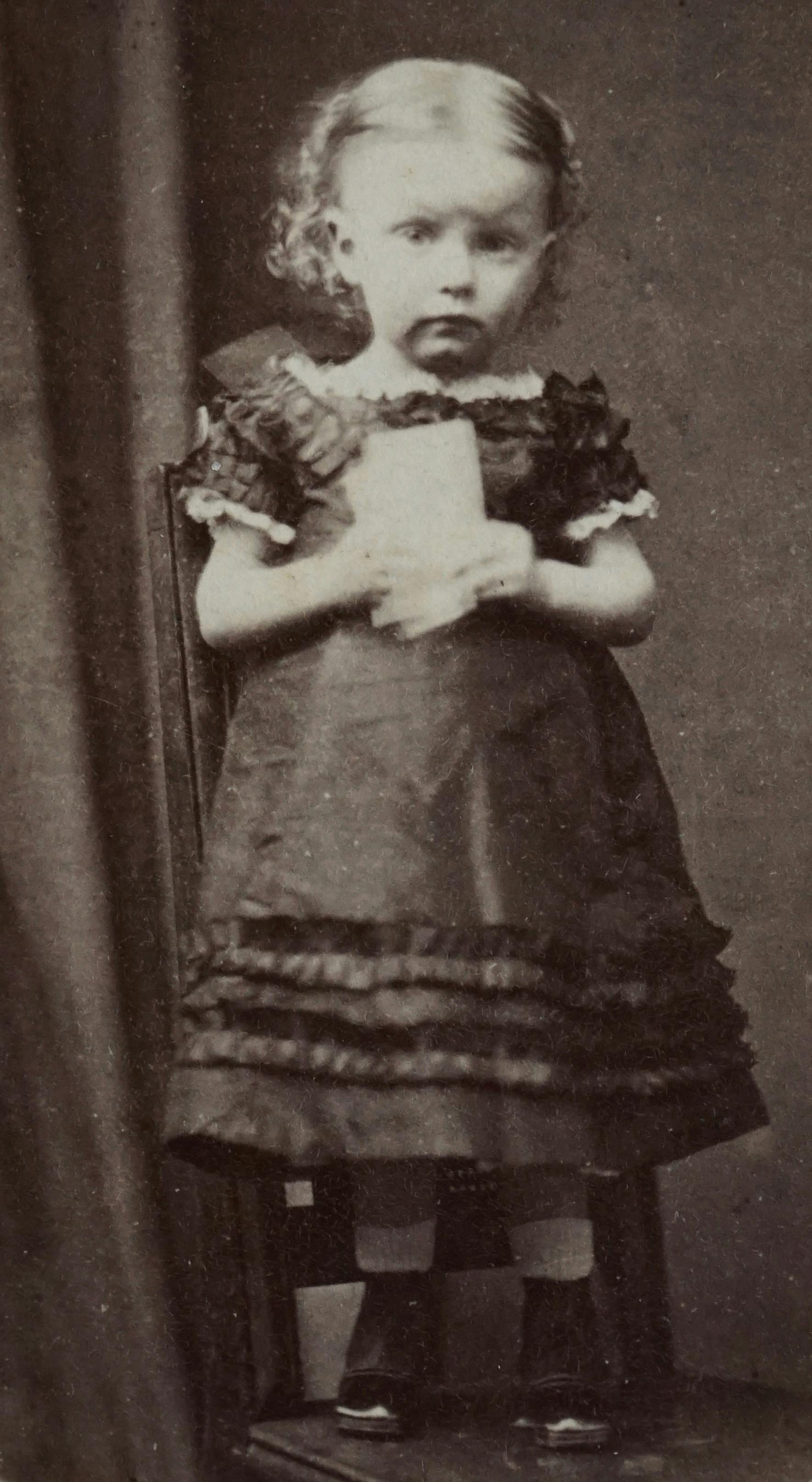 Antique black and white photo of a child in a vintage dress, standing.