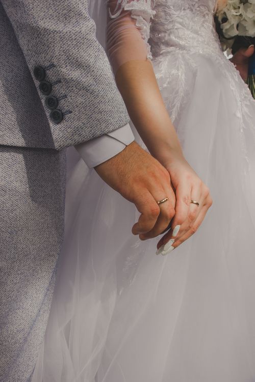 Free A beautiful close-up of a bride and groom holding hands, showcasing wedding rings and attire. Stock Photo