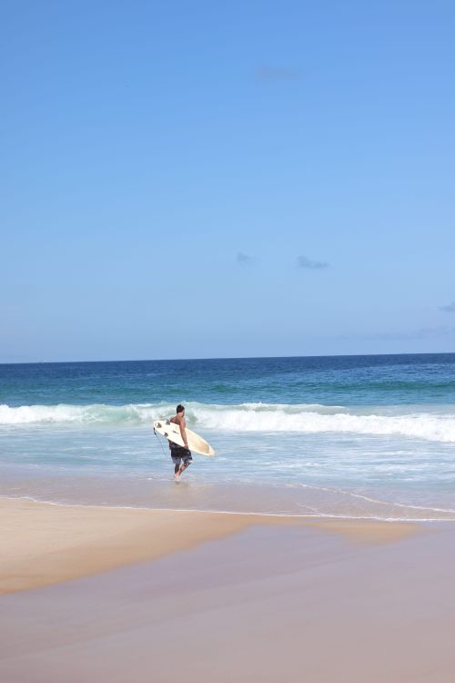 Free A surfer carrying a surfboard walks along Copacabana Beach in Rio de Janeiro, Brazil under clear blue skies. Stock Photo