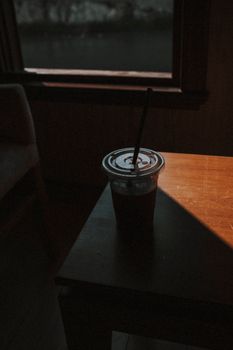 A moody indoor café scene featuring a plastic cup of iced coffee on a wooden table with shadow play.