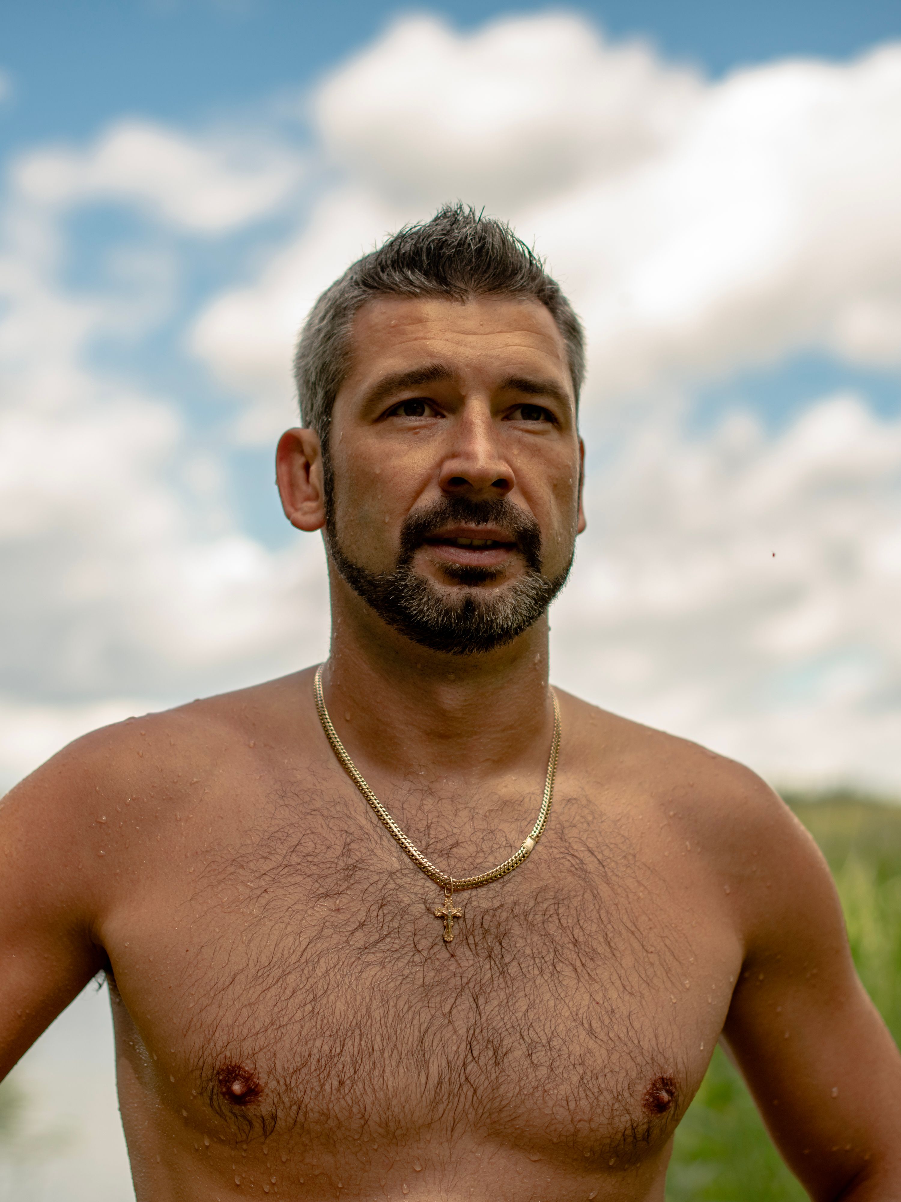 A fit man stands shirtless in nature, wearing a necklace and gazing into the distance.