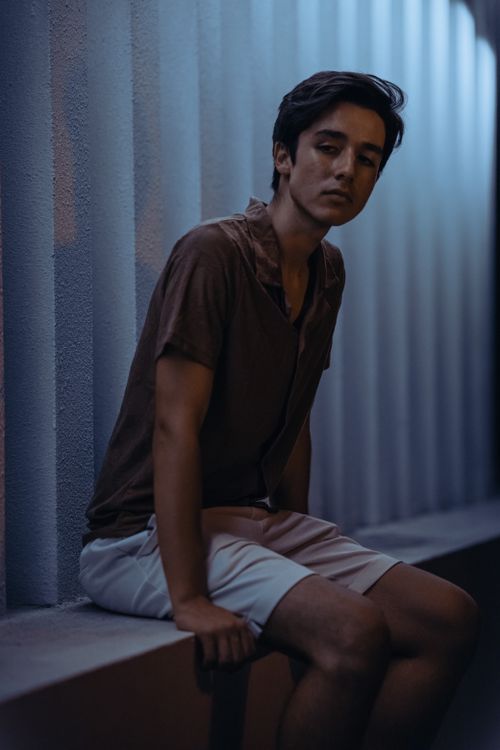 Free Young man sitting pensively on a bench by a textured wall in low light setting. Stock Photo