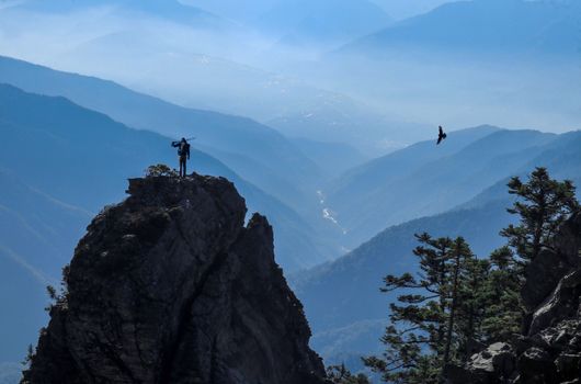 A lone hiker stands on a rocky peak, overlooking a vast mountain valley under a clear blue sky.