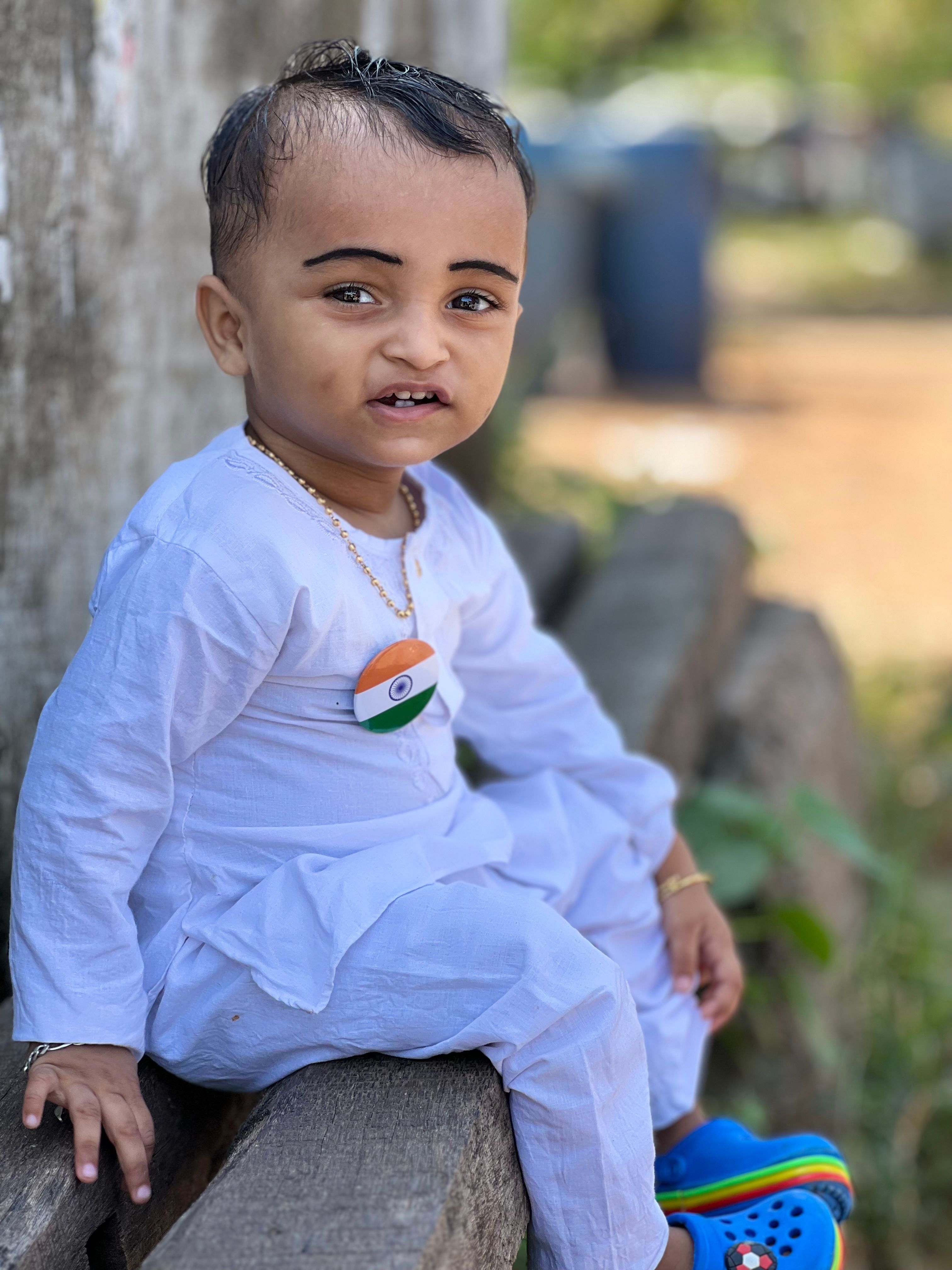 A young boy in Indian attire with a flag badge sitting outdoors with a natural background.