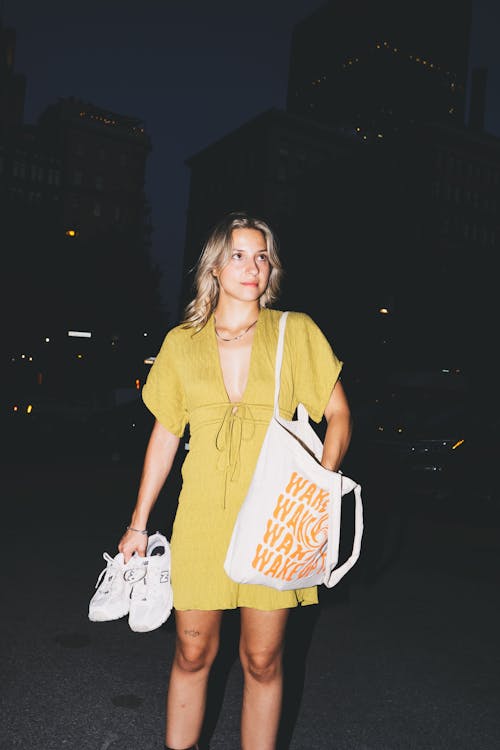 Young Woman in a Yellow Dress Holding Her Shoes and a Tote Bag Standing Outside at Night 