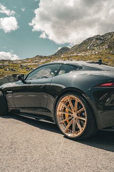 Black sports car with distinctive wheels on sunny mountain road, highlighting luxury and adventure.