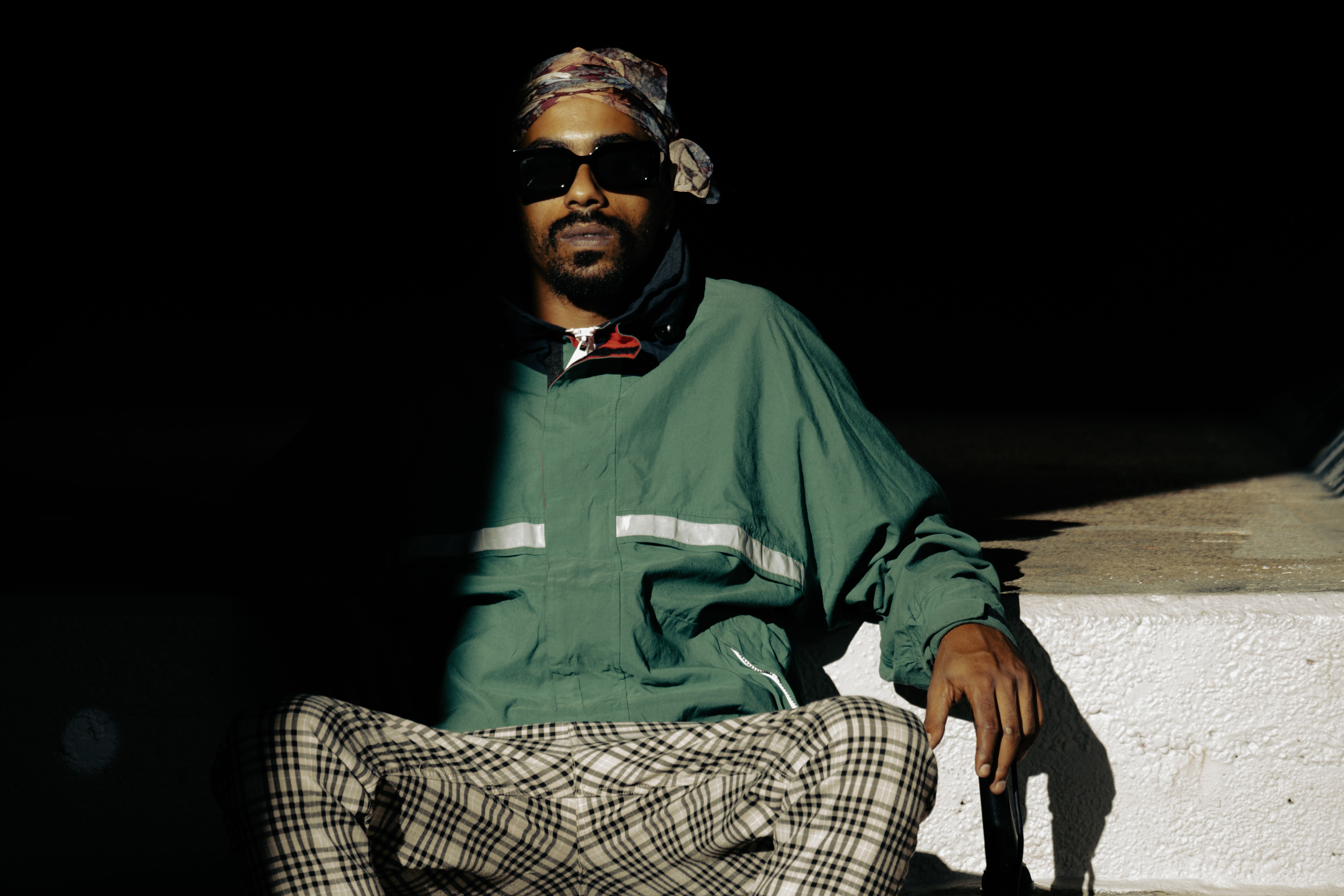 A stylish man wearing sunglasses sits on steps, partially in shadow, outdoors.