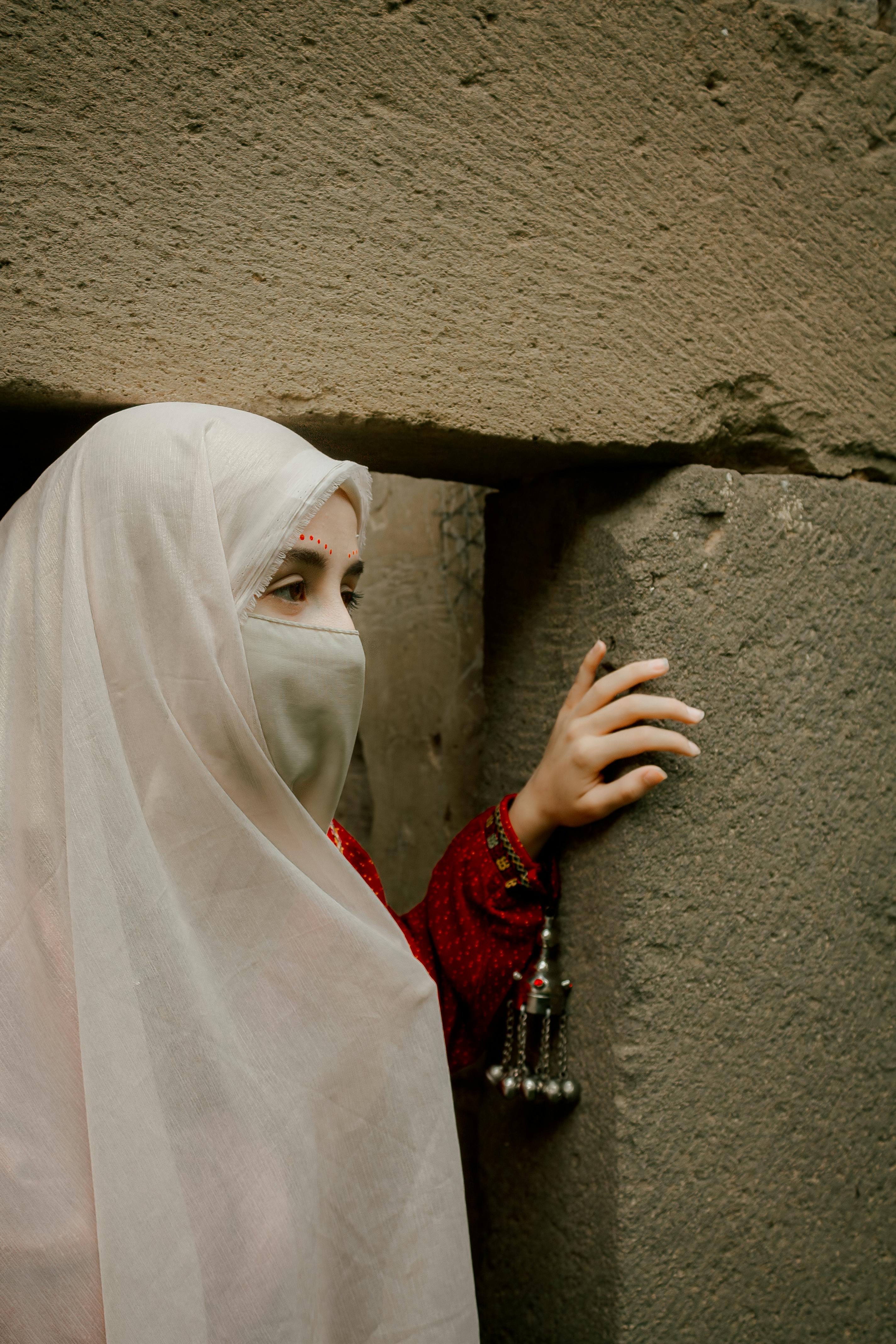 A woman wearing a white niqaab stands by a stone entrance, showcasing traditional attire.