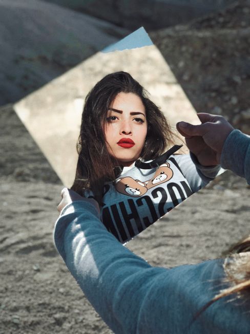 A woman holds a mirror outdoors, reflecting her face with striking red lips on a sunny day.