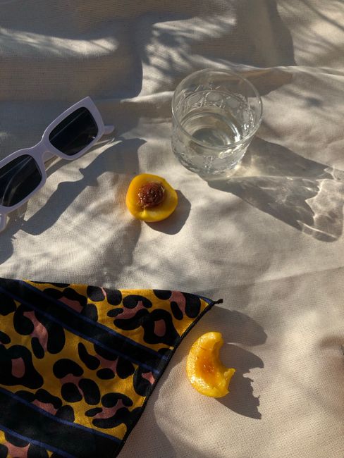 A sunlit composition featuring peaches, sunglasses, and a glass of water on patterned fabric.