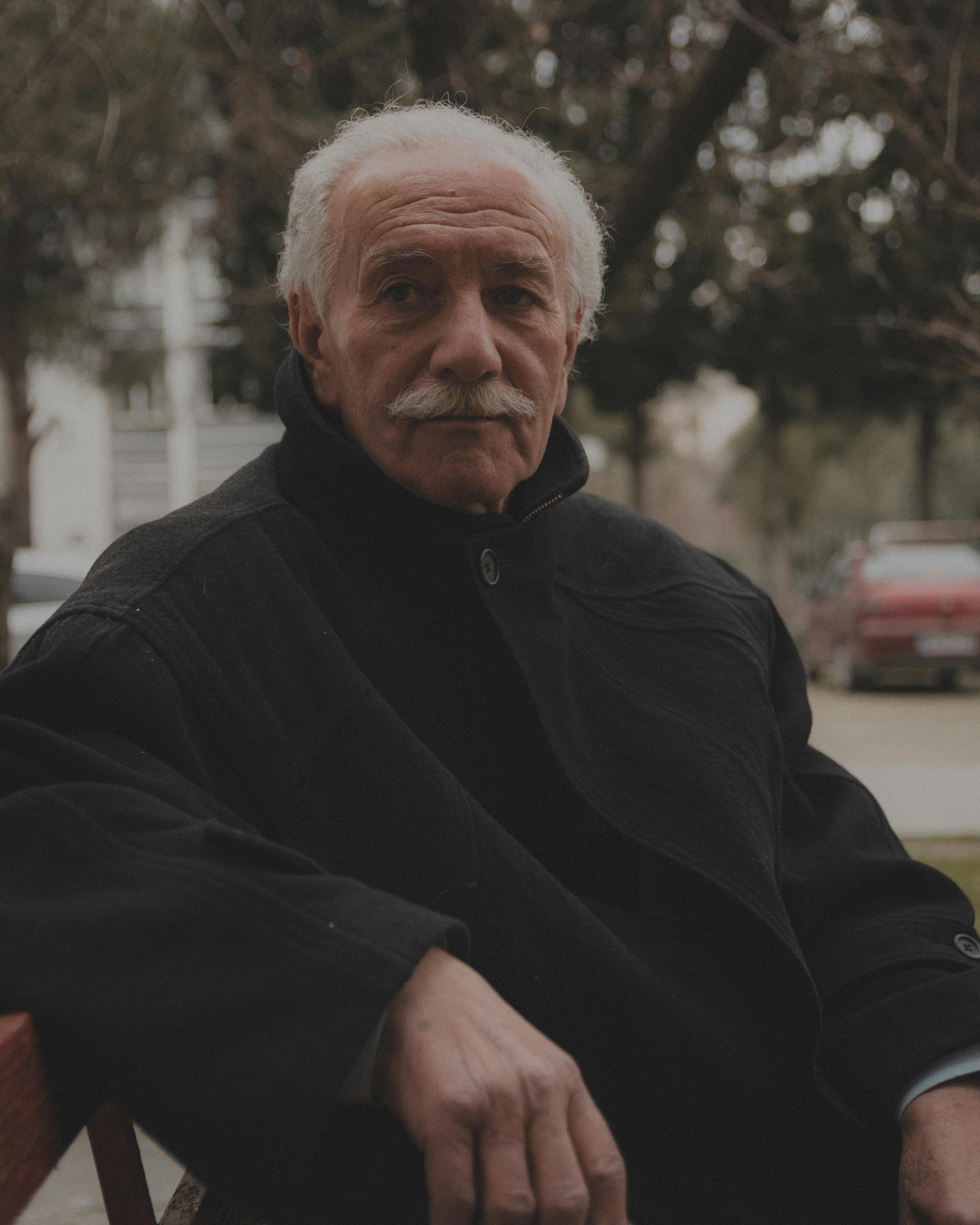 Portrait of an elderly man with gray hair sitting on a park bench outdoors.