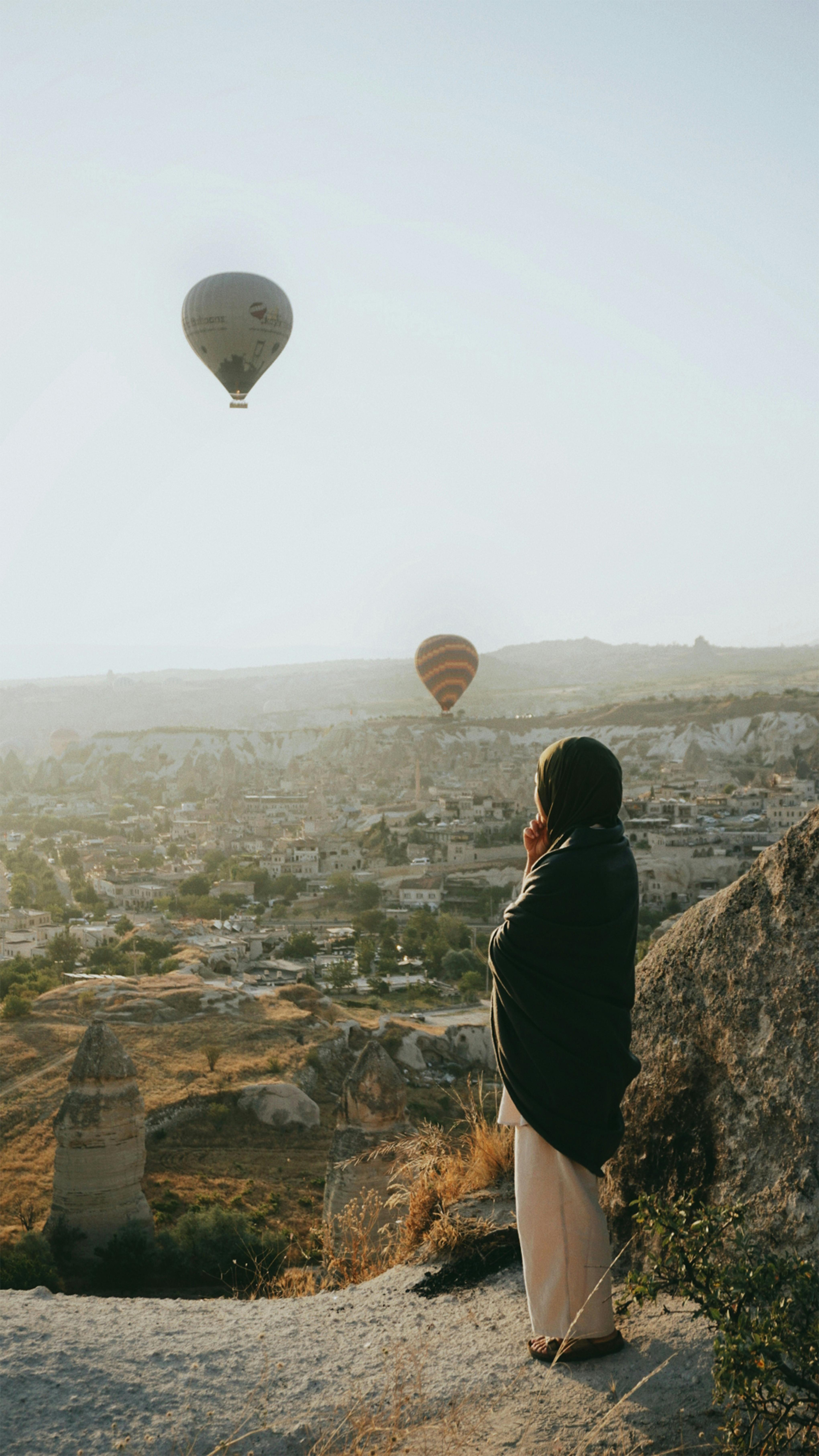 A woman stands on a cliff, observing hot air balloons over Cappadocia at sunset.
