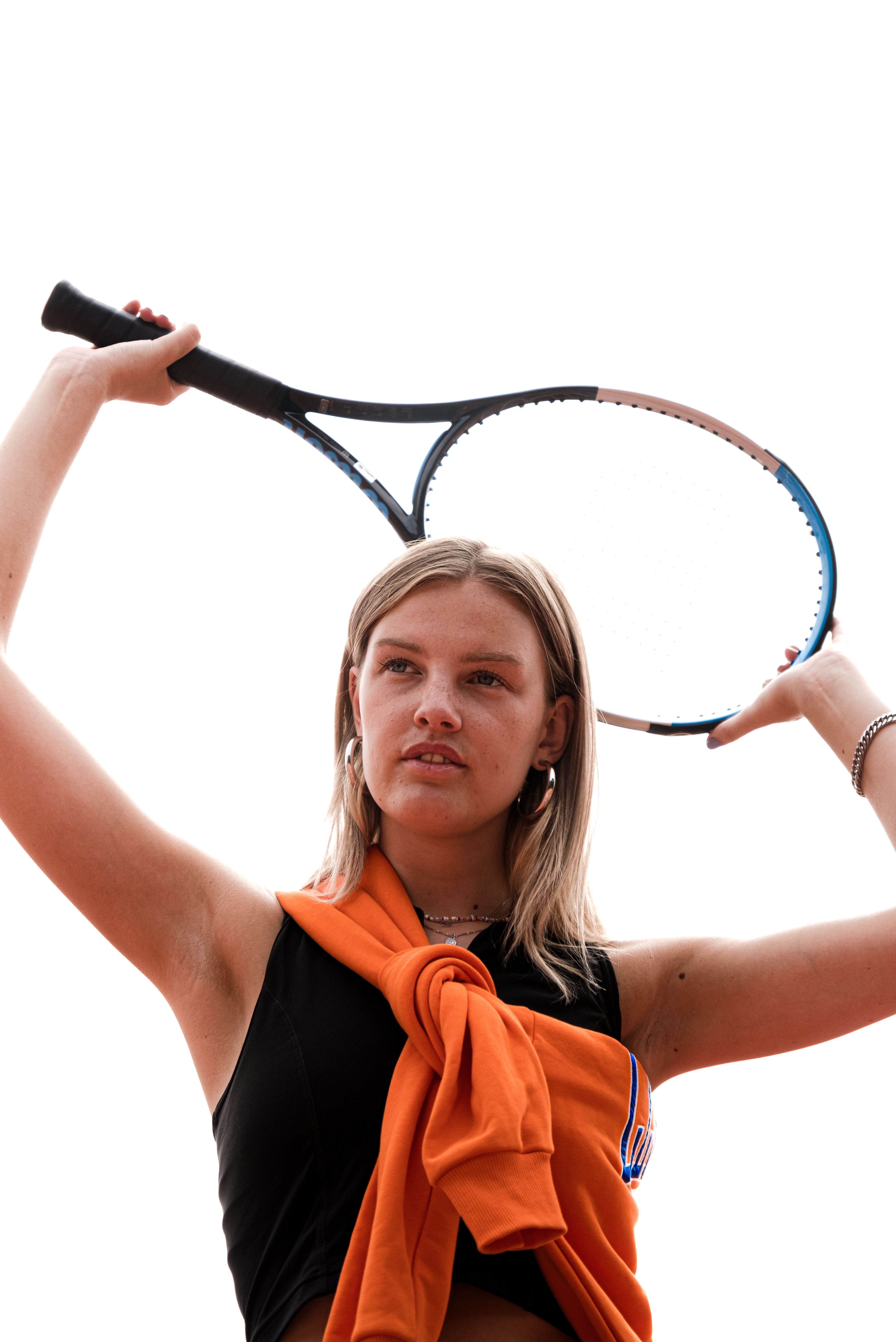 A confident young woman holding a tennis racket against a clear sky, showcasing athletic fashion.