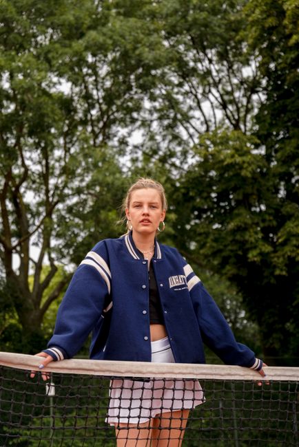 Confident woman in varsity jacket relaxes by tennis net outdoors.