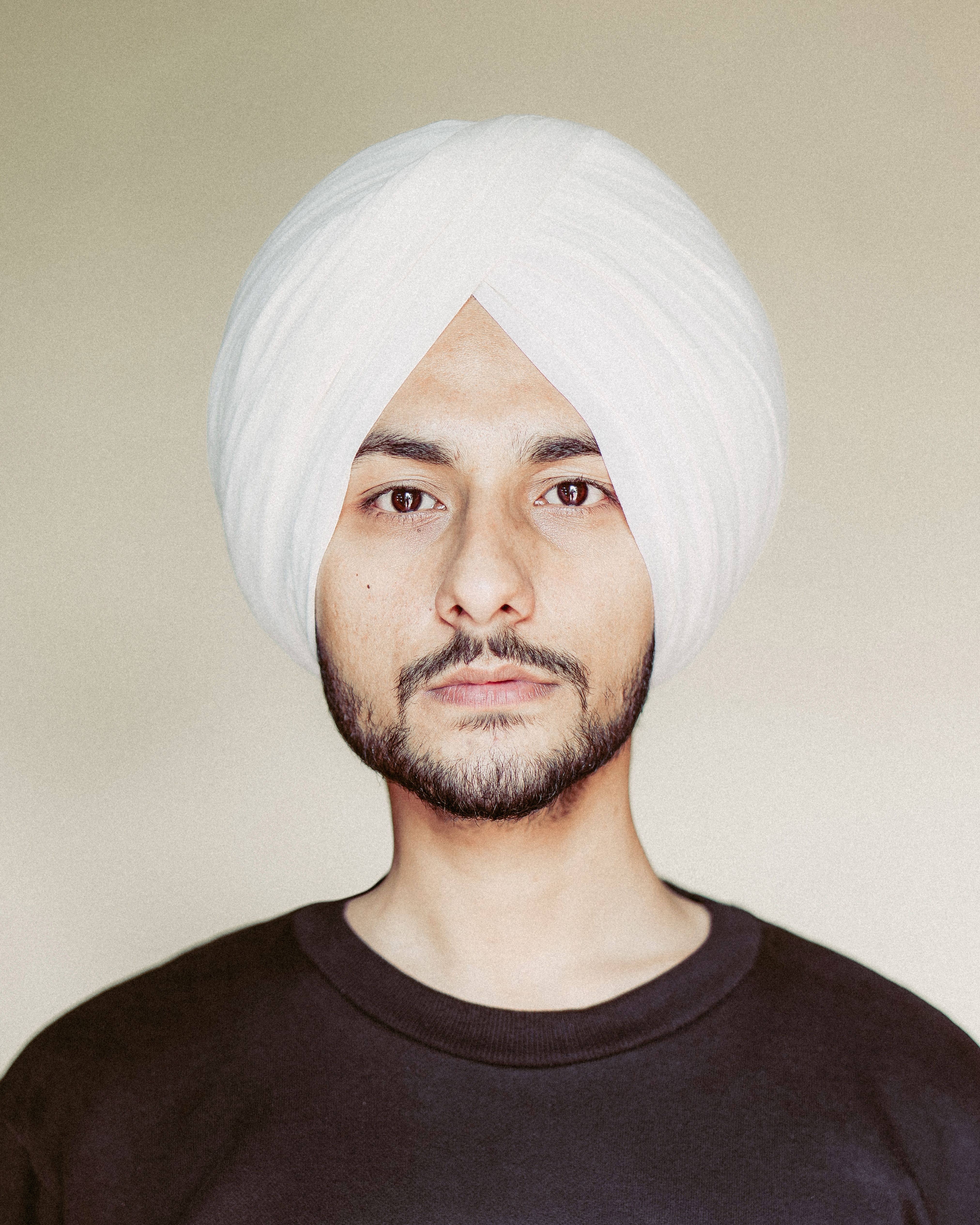 Close-up portrait of a man in a white turban, looking directly at the camera with a neutral background.