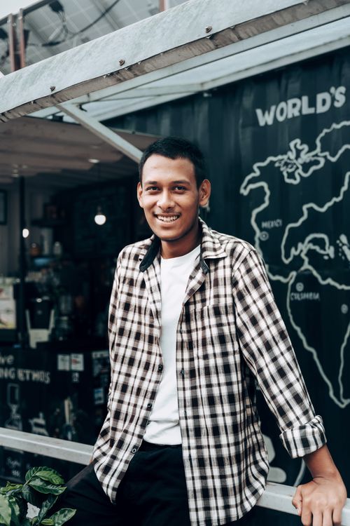 Free A cheerful man in a checkered shirt smiling while leaning against a railing at a café. Stock Photo