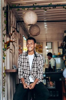 A cheerful young man in a checkered shirt sits in a cozy café with warm decor.