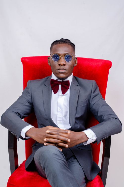 Free Stylish adult in a suit with bow tie, sunglasses, sitting on a classic red armchair with a serious expression. Stock Photo