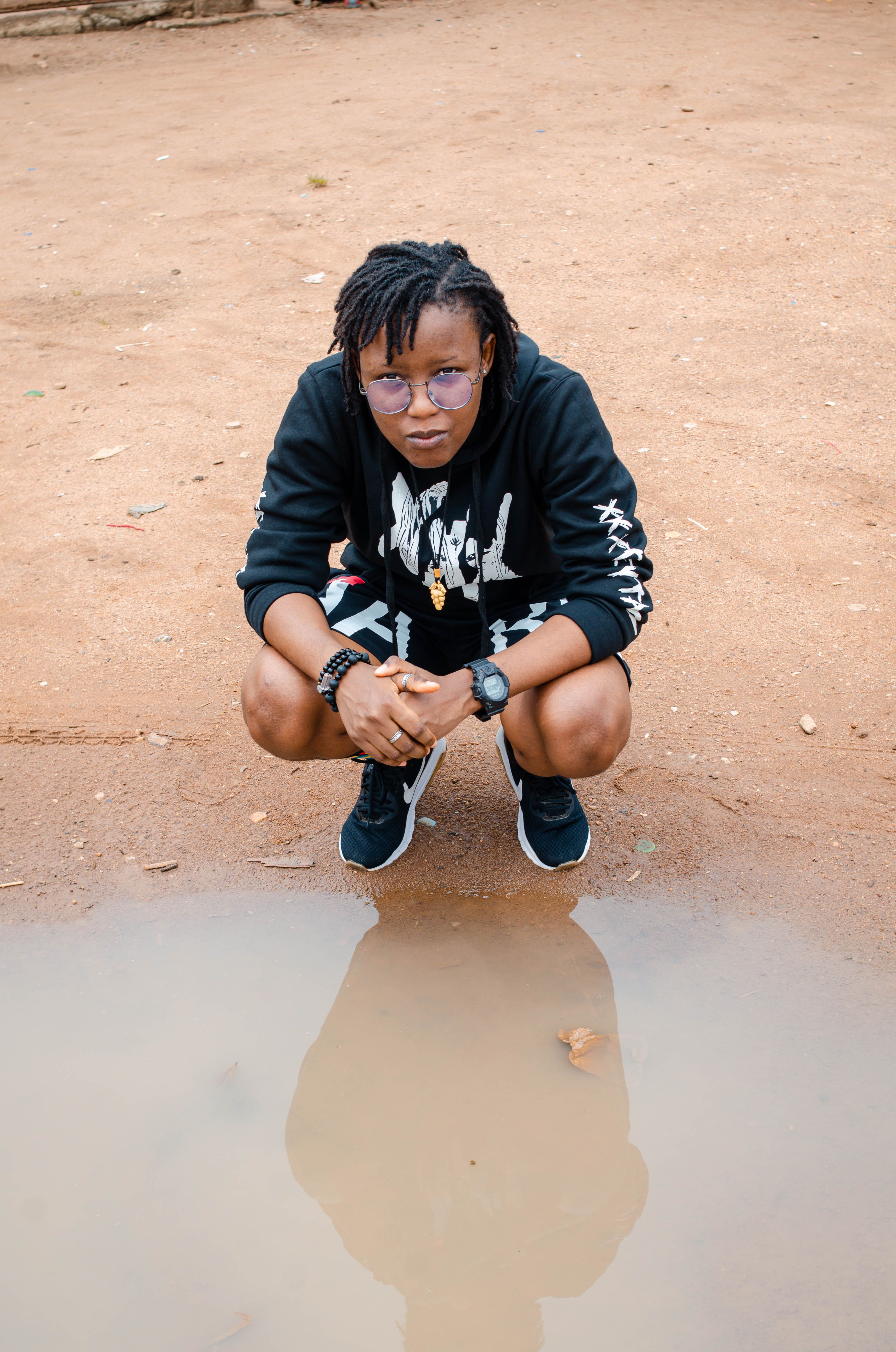 A young adult crouches on sandy ground overlooking a puddle, dressed casually in a hoodie and sneakers.