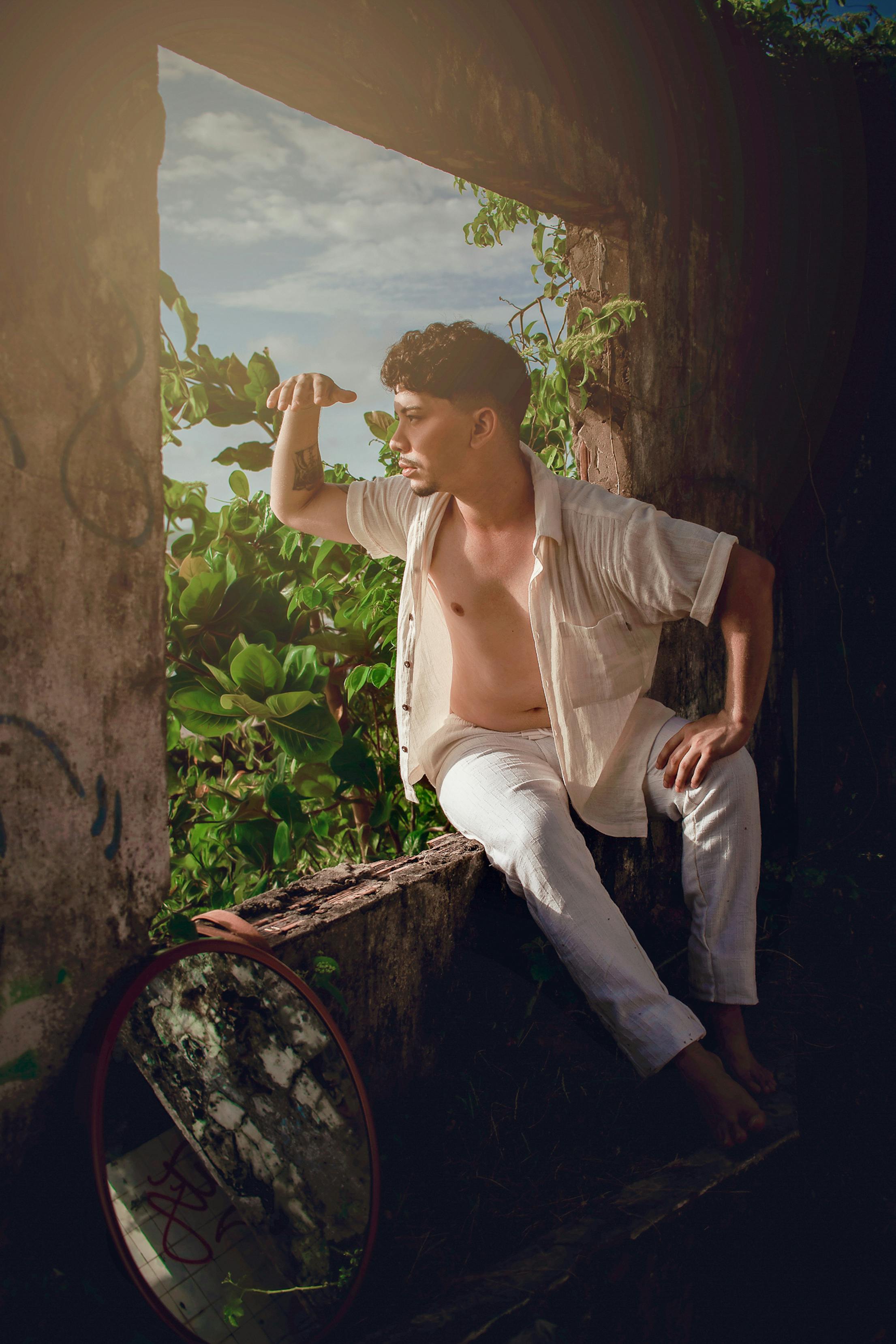 A fashionable young man in white attire sits by a window, exuding a relaxed summer vibe in São Luís.