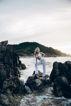 A woman in stylish attire poses confidently on rocky seashore under bright daylight.