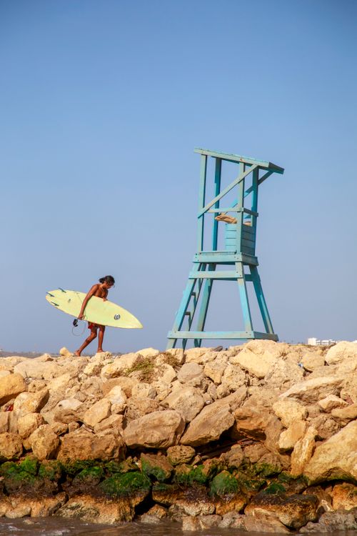 Free A surfer carrying a board walks beside a blue lifeguard tower on a rocky beach in Barranquilla, Colombia. Stock Photo