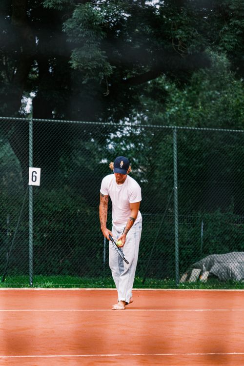 Free Adult playing tennis, preparing to serve on a clay court surrounded by trees. Stock Photo