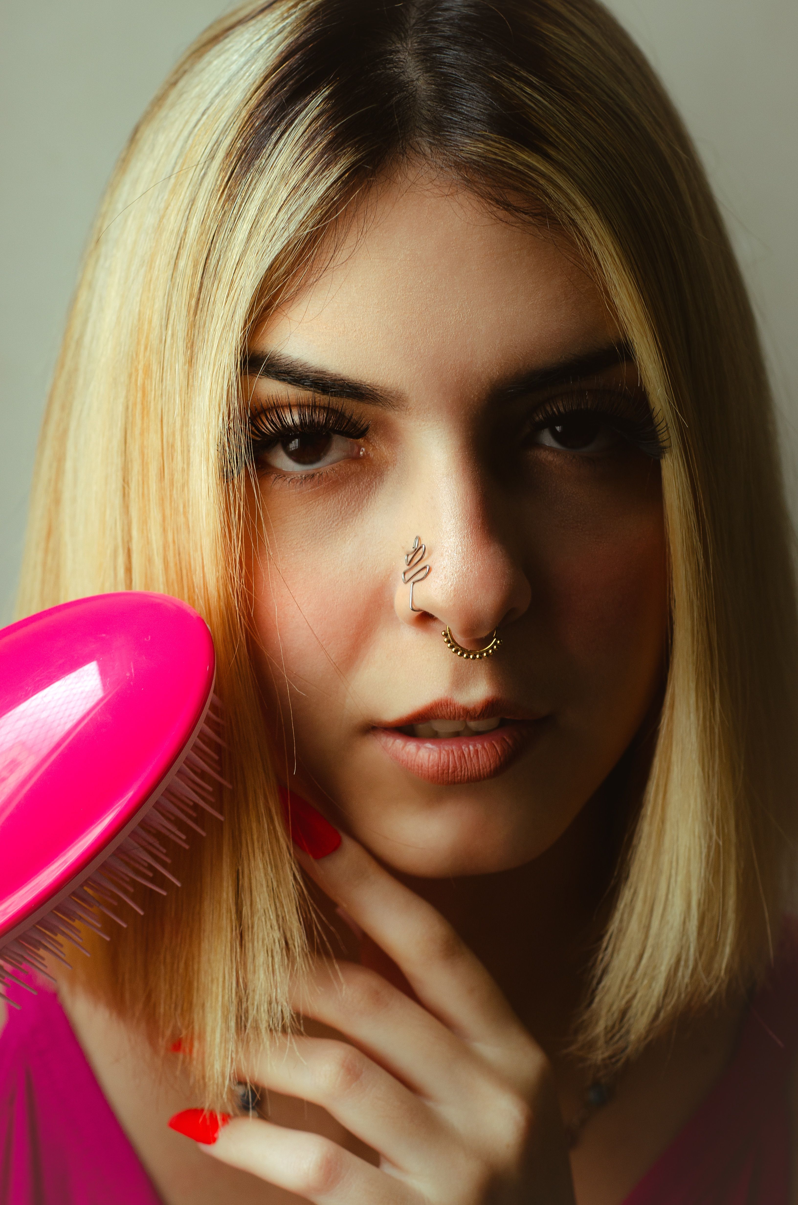 Close-up portrait of a blonde woman with nose piercing holding a pink comb, exuding elegance.