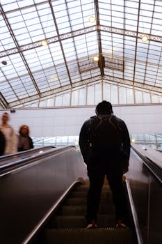 A man with a backpack ascends an escalator indoors, under a glass ceiling.