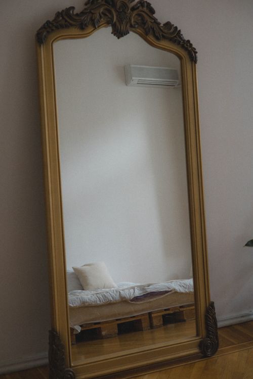Free A large ornate mirror reflects a minimalist bedroom with a bed on wooden pallets, showcasing stylish interior design. Stock Photo