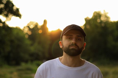 Free A bearded man in a cap stands outdoors at sunset, bathed in warm sunlight. Stock Photo