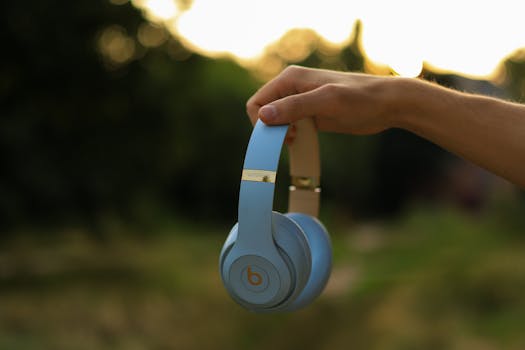 A hand holding blue wireless headphones outdoors during sunset, with a blurred natural background.