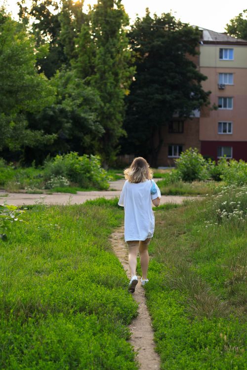 Free A blonde woman walks along a narrow path in a grassy urban area, surrounded by trees and buildings. Stock Photo