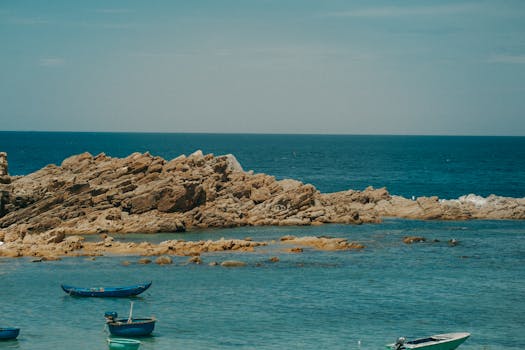 Tranquil coastal scene with boats floating by rugged rocks under a bright blue sky.