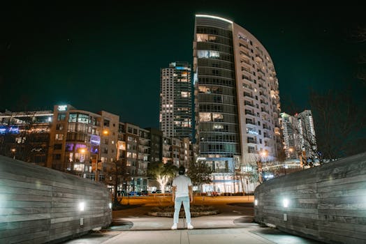 夜幕下的现代都市，一个孤独的身影面向摩天大楼，凸显出城市生活的寂寥