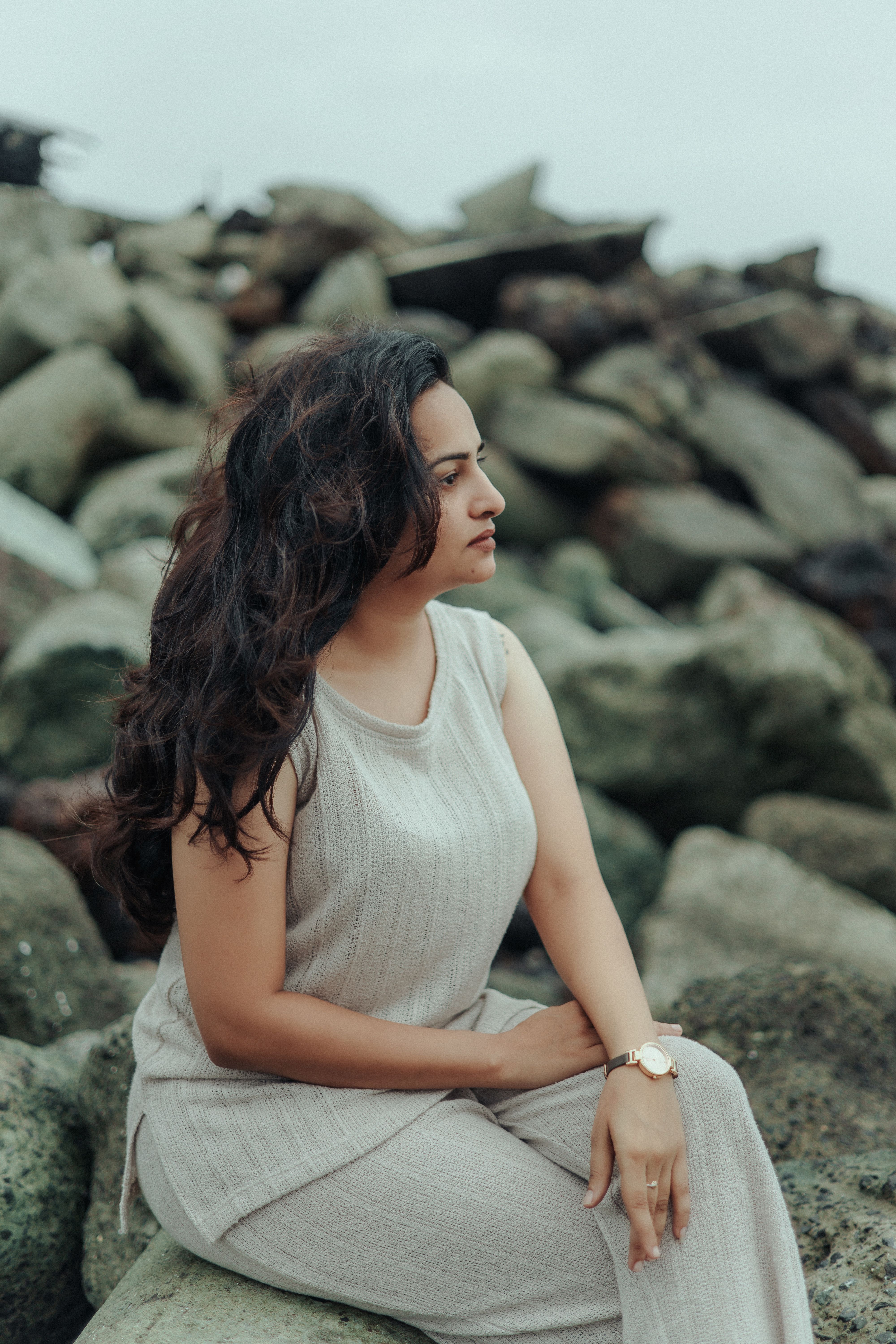 A serene woman in a beige outfit poses thoughtfully on a rocky breakwater.