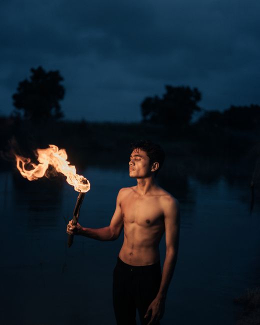 A topless young man holds a flaming torch by a lake at night, creating a dramatic portrait.