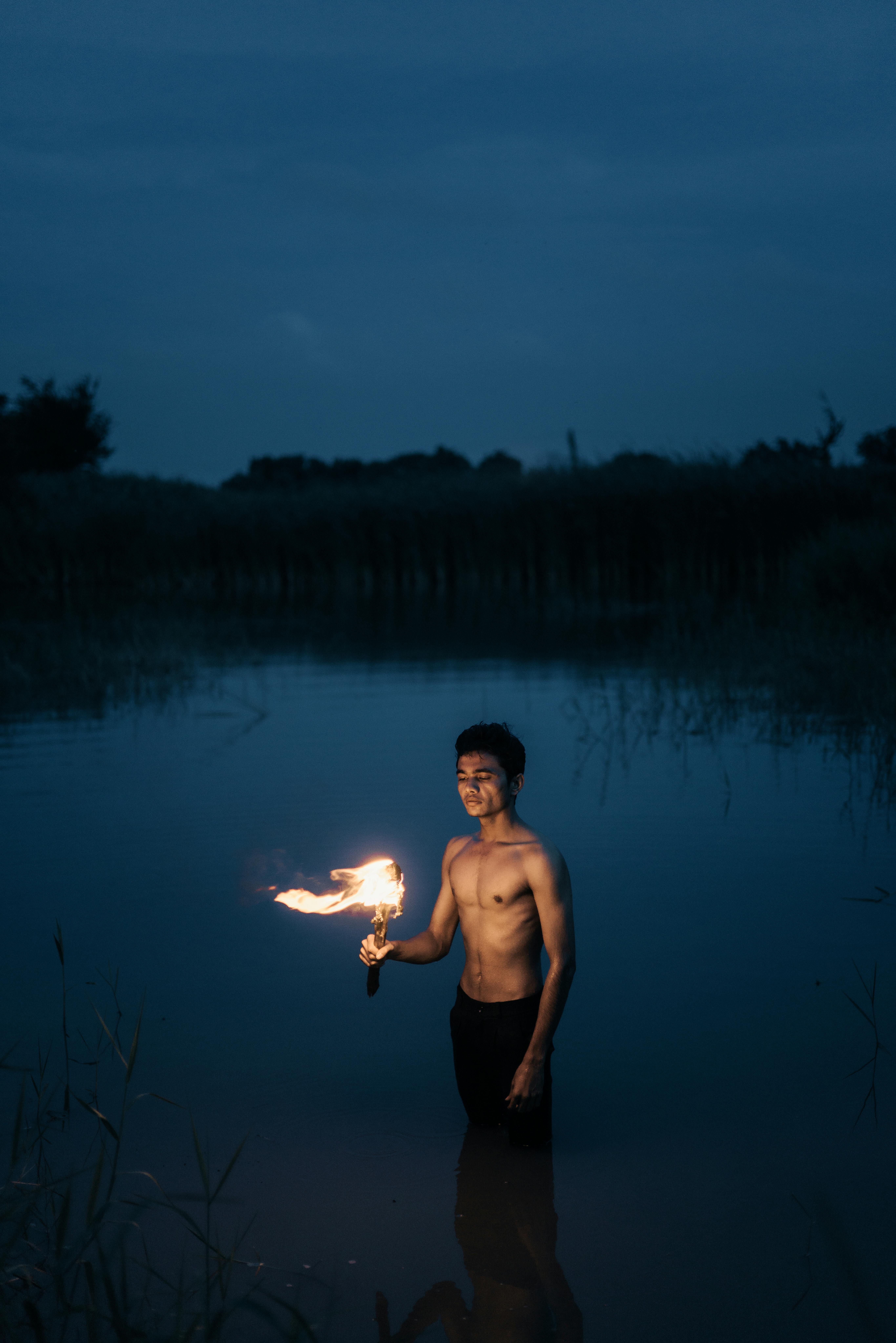 Topless man stands in water at night, holding a flaming torch, creating a dramatic scene.
