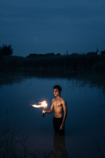 Topless man stands in water at night, holding a flaming torch, creating a dramatic scene.