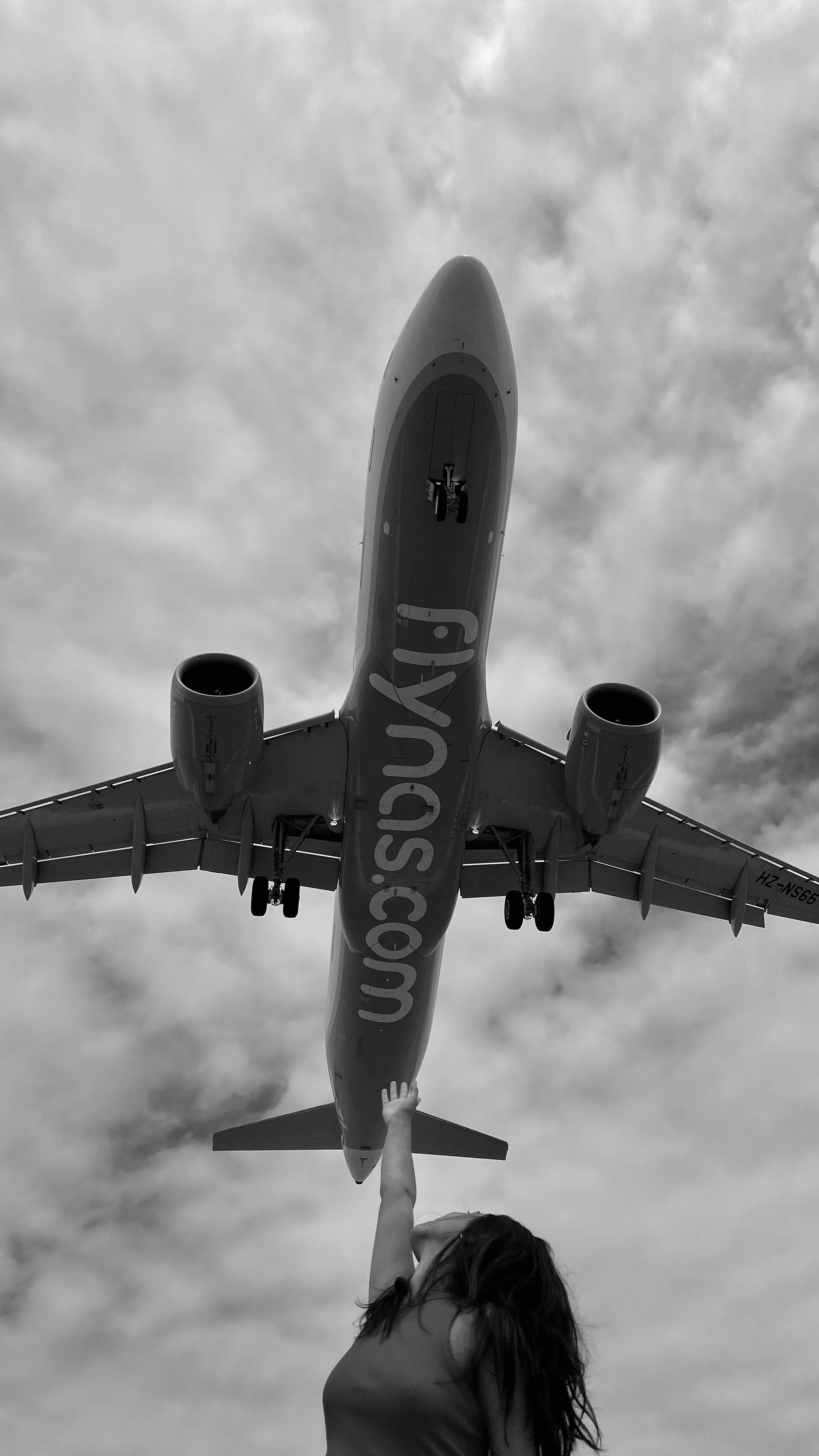 A woman reaches up towards a low flying airplane captured in black and white.