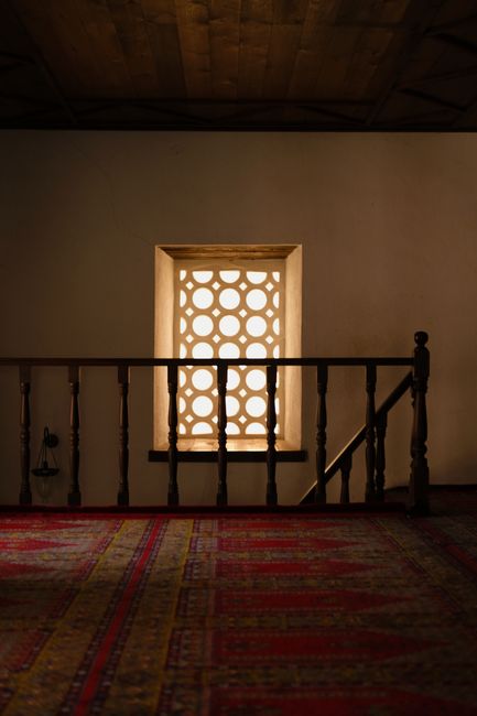 A dimly lit corridor featuring an ornate window and traditional decor, casting patterns on the floor.